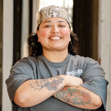 Smiling person with tattoos and a camouflage cap, arms crossed confidently.
