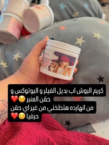 A hand holding a cream jar with a woman's image, surrounded by other jars on a star-patterned blanket.