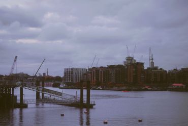 Cloudy riverside cityscape with cranes and buildings along the water.