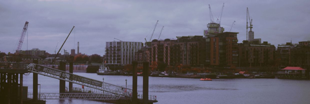Urban waterfront with cranes, buildings, and a dock under cloudy skies.