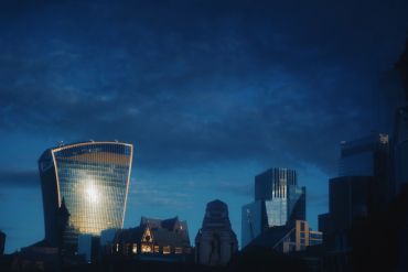 City skyline at dusk with illuminated modern buildings under a cloudy sky.