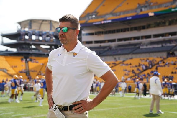 A man in a white polo with sunglasses stands on a football field.