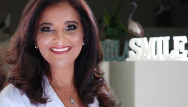 A smiling woman with dark hair and makeup in front of a 'SMILE' sign.