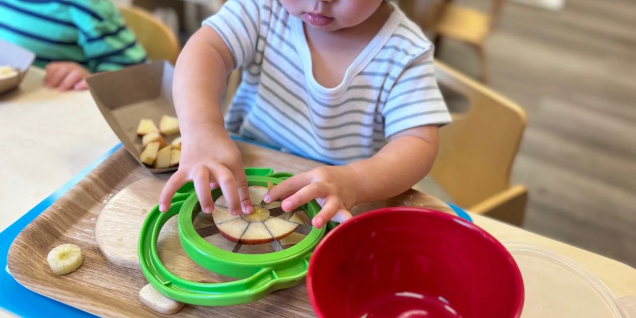 A child slices an apple using a green apple slicer on a wooden tray.