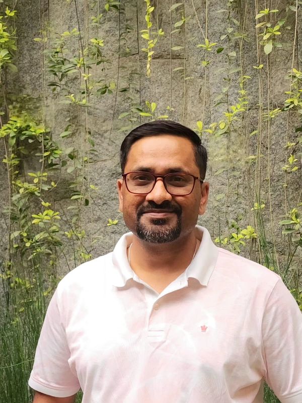 A man with glasses in a white polo shirt stands outdoors against a green vine-covered wall.