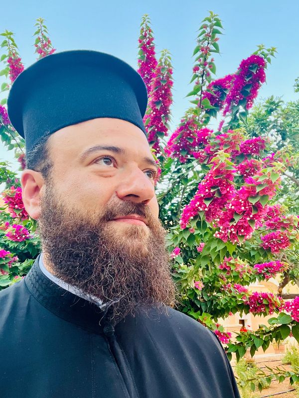 Bearded man in religious attire stands before bright pink flowers under a clear blue sky.