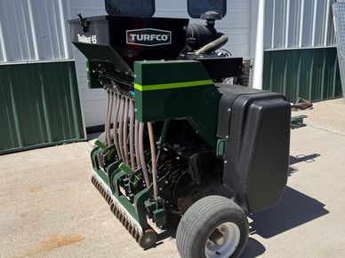 A green Turfco machine with tubes and wheels on a concrete surface outside a building.