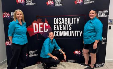 Three people in teal shirts pose in front of a Disability Events Community banner.