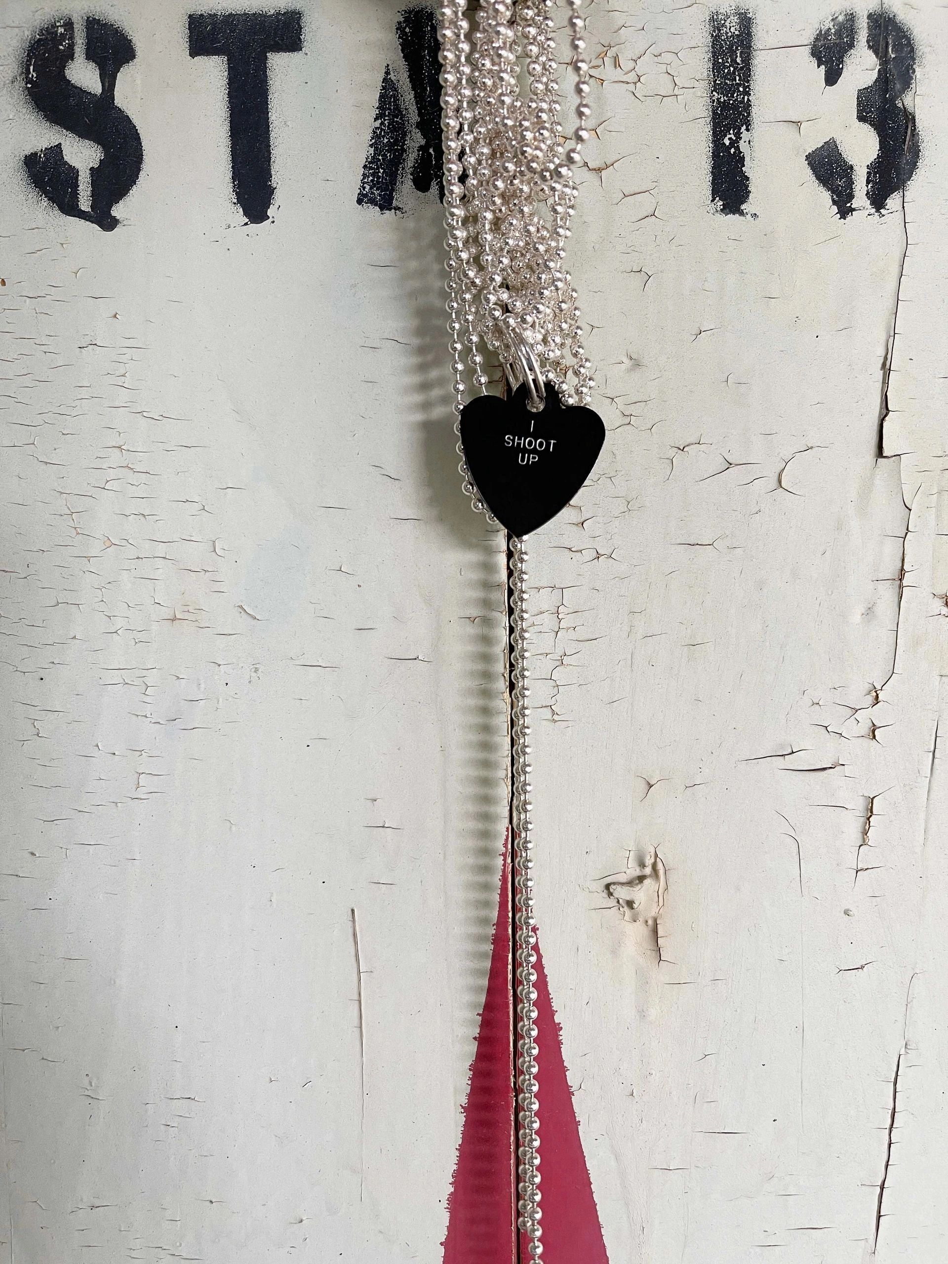 Silver bead necklace with a black heart pendant saying 'SHOOT UP'.