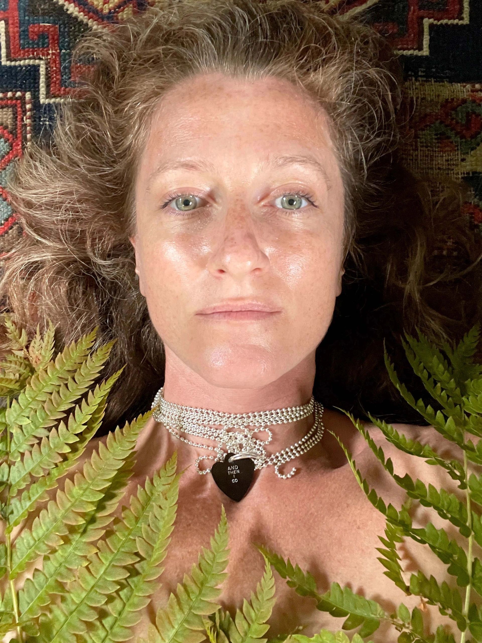 Woman lies on a patterned surface surrounded by green ferns wearing a silver necklace.