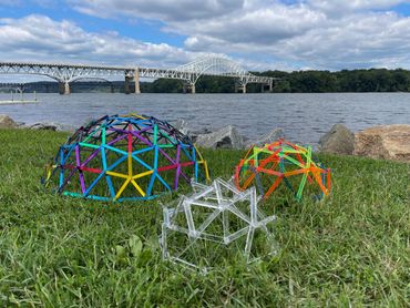 Colorful geometric structures on grass near a river with a bridge in the background.