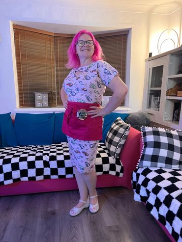 Woman with bright pink hair wearing a patterned dress and red apron in a cozy living room.