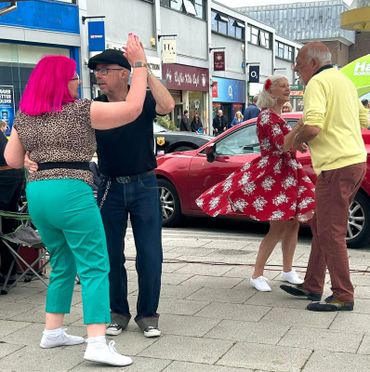 Two couples dance joyfully on a city street with colorful outfits and lively expressions.