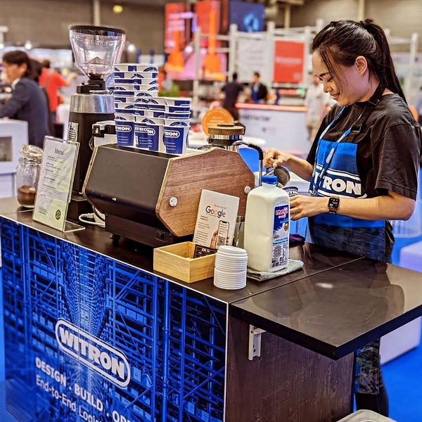 Barista preparing coffee at a WITRON branded coffee stall in a busy indoor event.