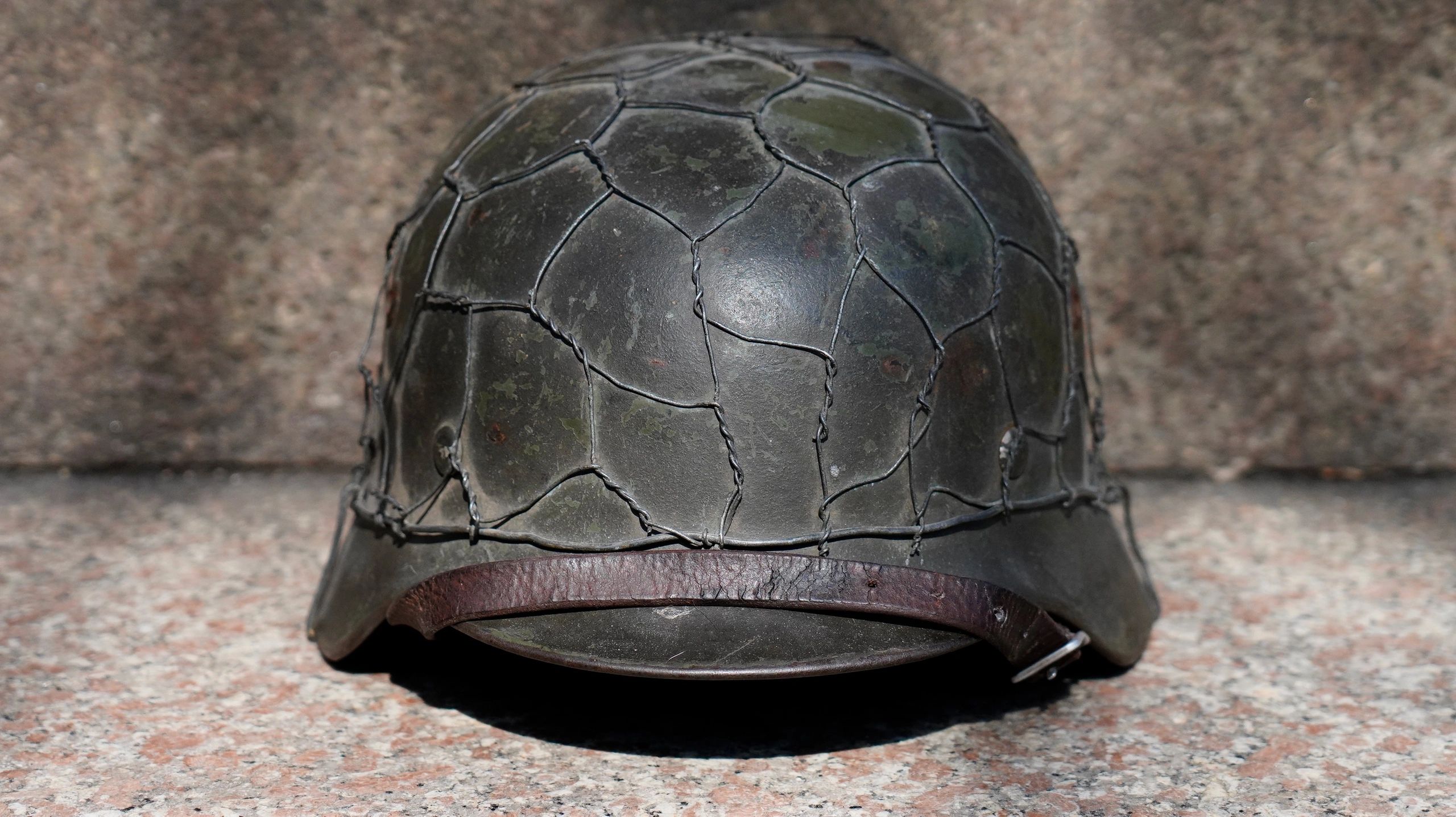 Old military helmet with wire netting and leather strap on a stone surface.