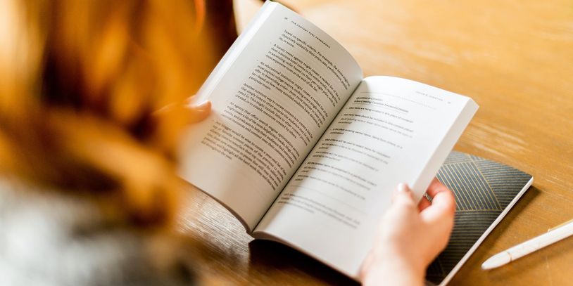 Person reading a book at a wooden table with a notebook and pen.
