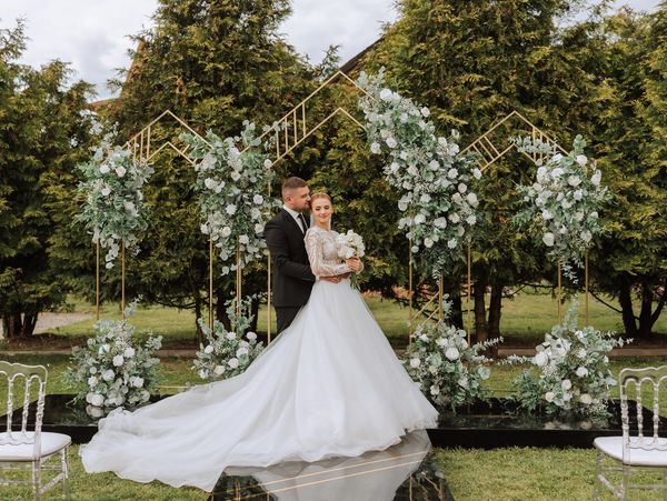 A groom embraces his bride in a white gown and holding flowers, under a geometric floral arch.