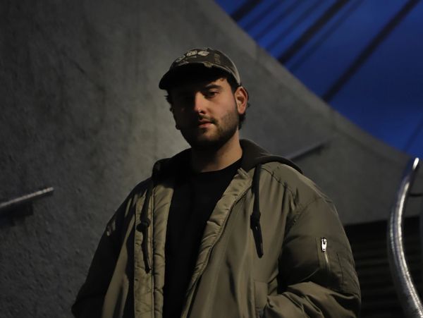 Man in a green jacket and cap standing in a dimly lit urban setting at night.