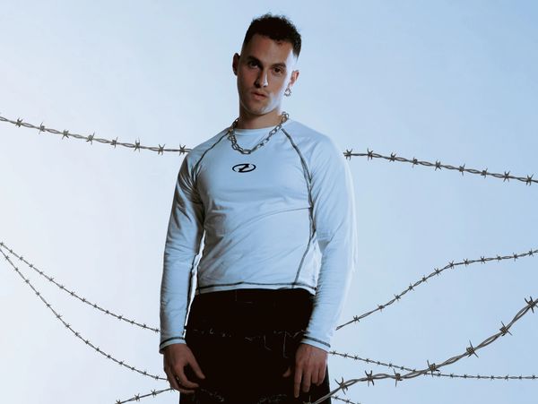 Young man in white shirt and chain necklace stands against a barbed wire backdrop.