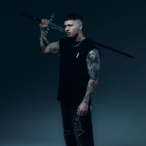 Tattooed man holding a large sword over his shoulder, wearing black clothing.