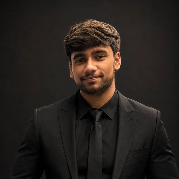 Young man in a black suit and tie smiling against a dark background.