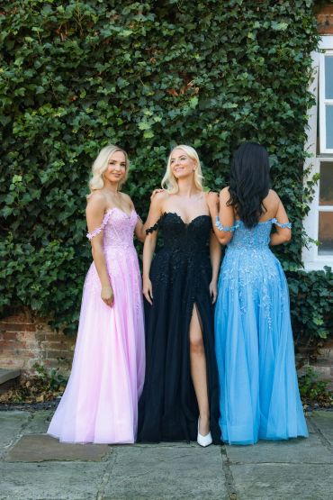 Three women in elegant dresses pose outdoors against ivy.
