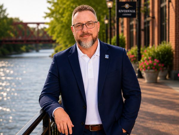 Man in a navy blazer and white shirt standing by a riverside railing.