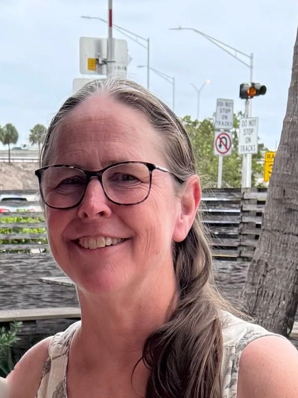 Smiling woman with glasses and long hair outdoors near a palm tree.