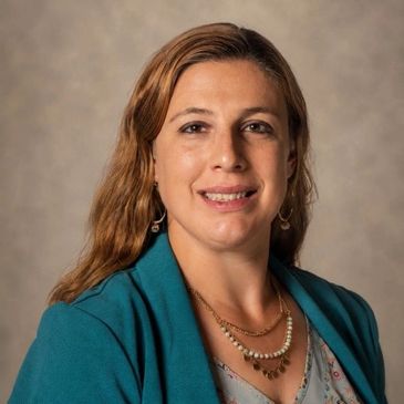 Professional woman smiling, wearing teal jacket and layered necklaces.