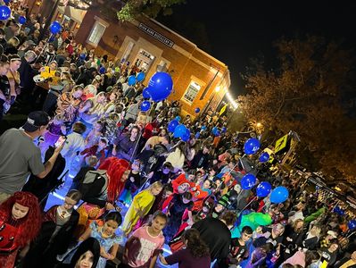 Children and families in costumes gather outdoors at night for a festive event with blue balloons.
