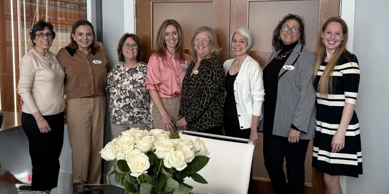 Group of eight smiling women posing indoors with white roses in front.