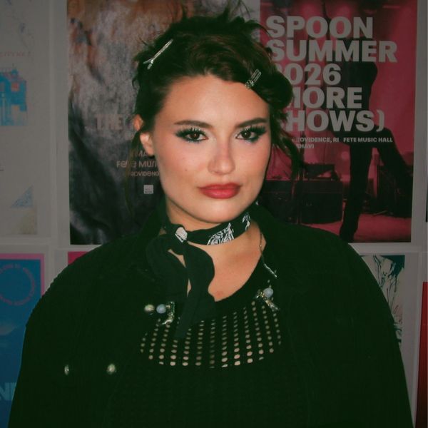 Woman with dark hair and bold makeup in front of music posters.