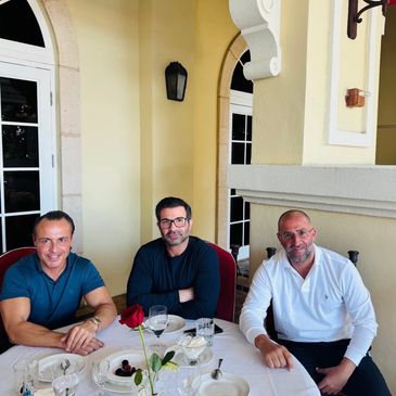 Three men sitting at a round dining table with a rose centerpiece, smiling.