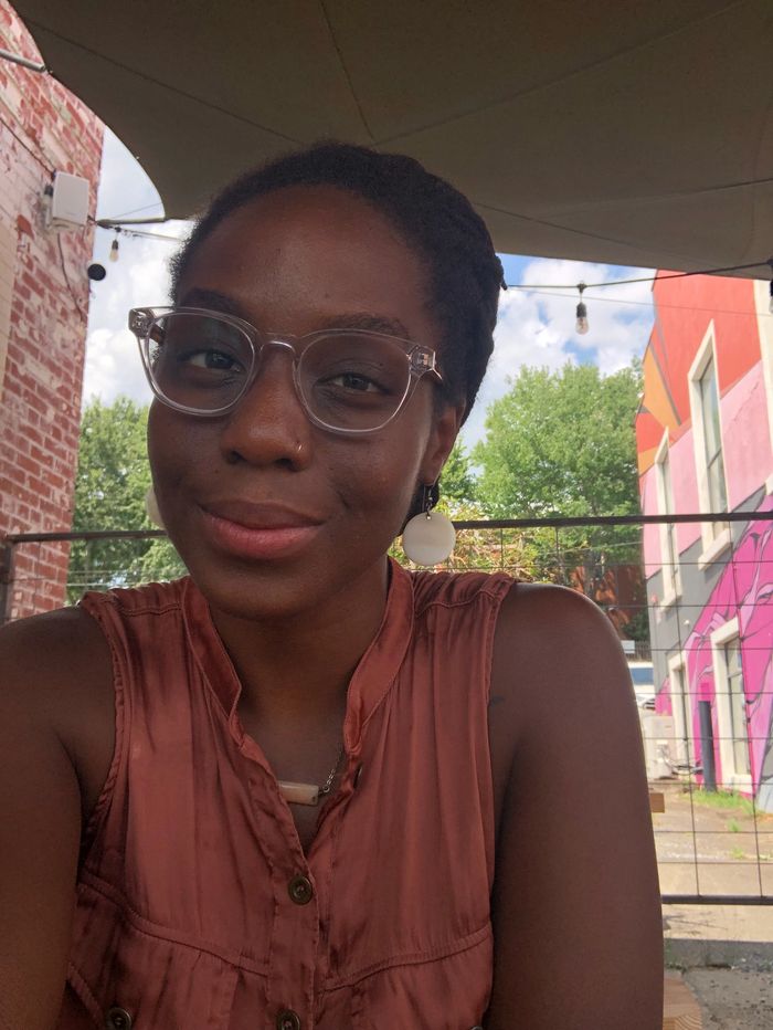 Aqiyla smiles with glasses on with the Carolina blue sky and a colorful mural behind her.