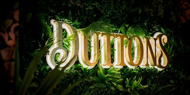 Illuminated sign reading 'JUTTONS' surrounded by green plants.