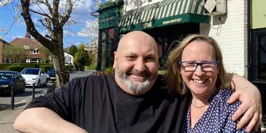 Smiling couple posing outside a restaurant with blooming tree nearby.