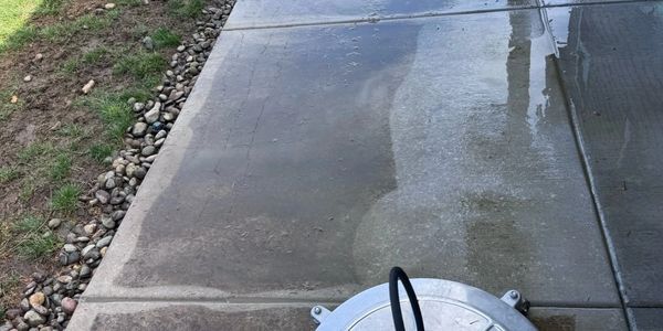 Pressure washer cleaning a concrete driveway near grass and rocks.