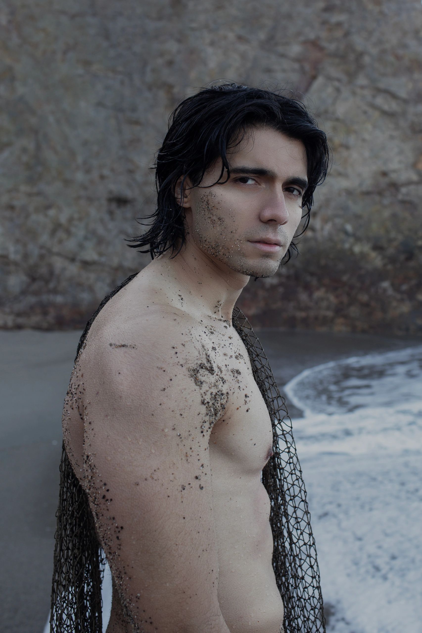 A shirtless man with wet hair stands on a beach, covered in sand and holding a net.