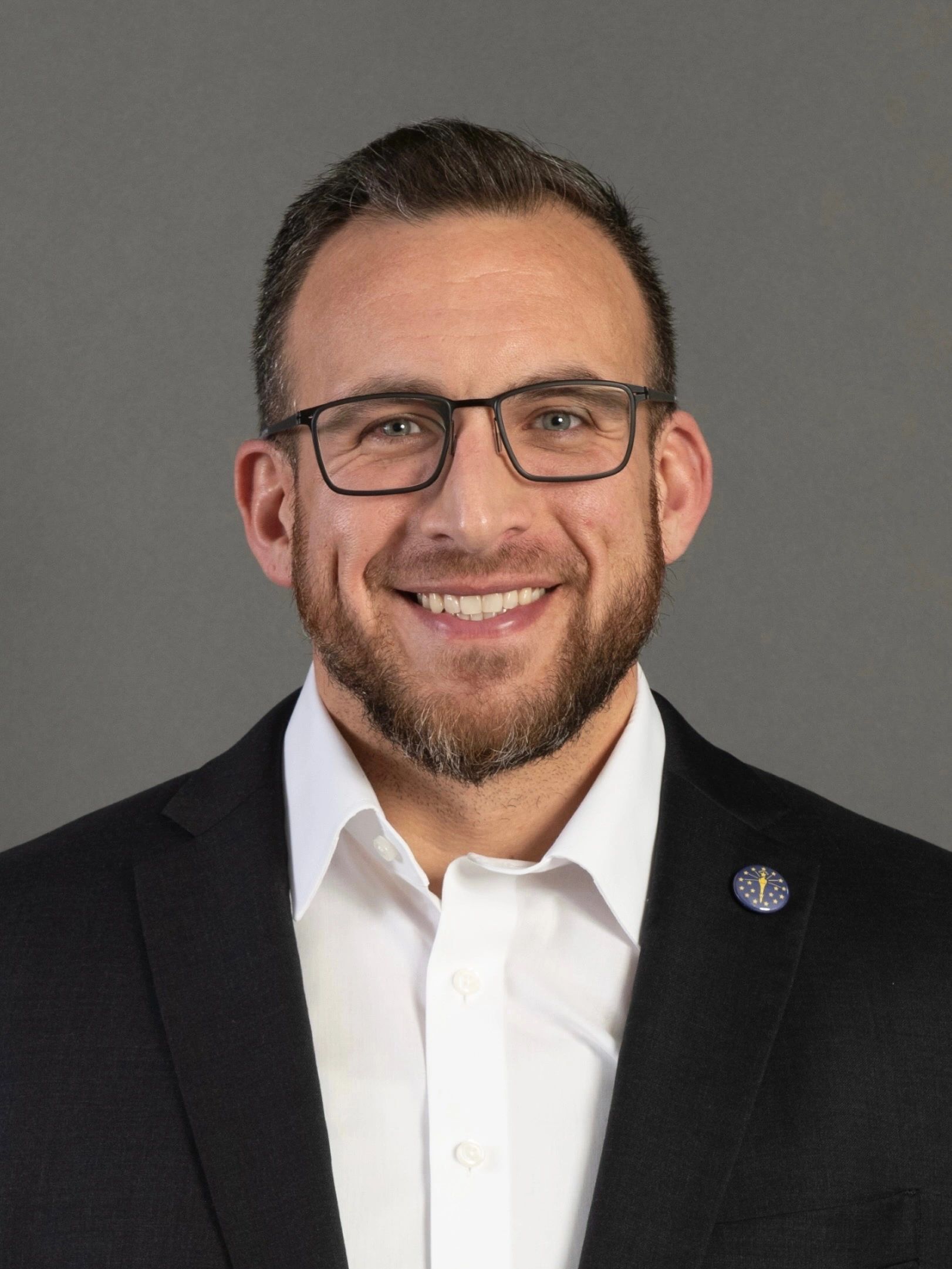 Smiling man wearing glasses and a suit jacket with a lapel pin.