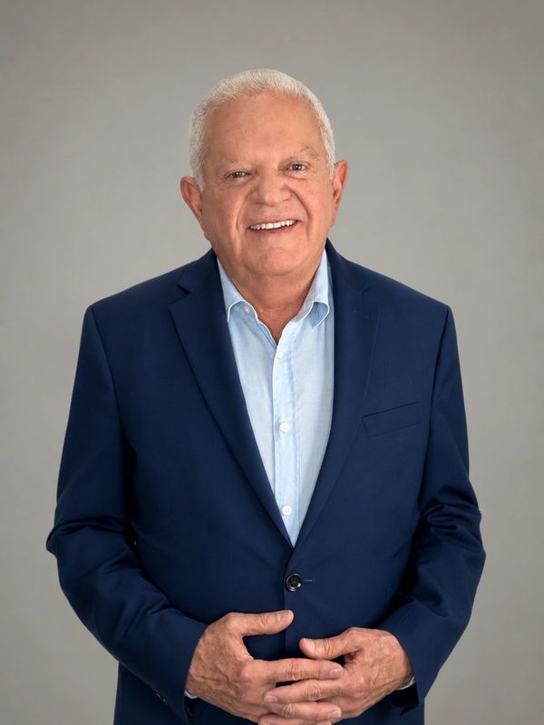 Smiling elderly man in a blue suit and light blue shirt against a plain background.