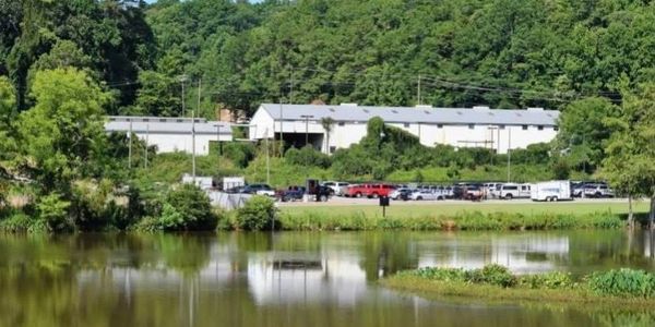 A serene pond with a white industrial building and parked vehicles surrounded by trees.