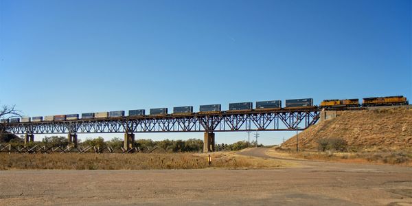 A long freight train crosses a tall metal bridge under a clear blue sky.