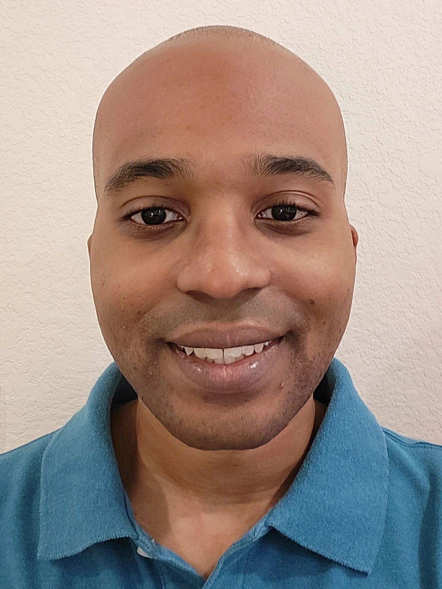 Smiling bald man wearing a blue garment against a textured wall.