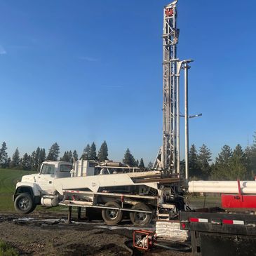 White drilling rig truck operating in a green field under clear blue sky.