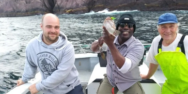 three people in a boat off shore. The person in the middle smiling with excitement holding a cod fis