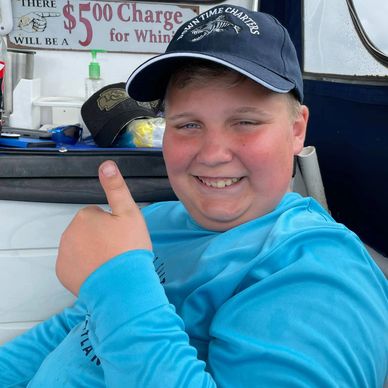 Smiling boy in blue fishing shirt and navy cap gives thumbs up on boat.