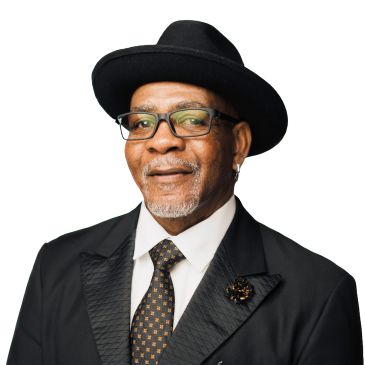 Elderly man in a black hat and suit with glasses, smiling.