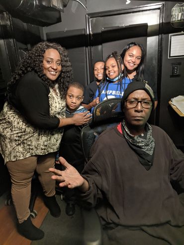 A group of five people posing happily indoors with one person seated in front.