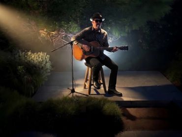 Musician playing guitar on a small outdoor stage at night.