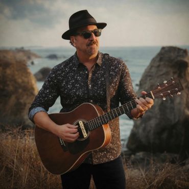 Man wearing a hat playing guitar by the seaside during sunset.